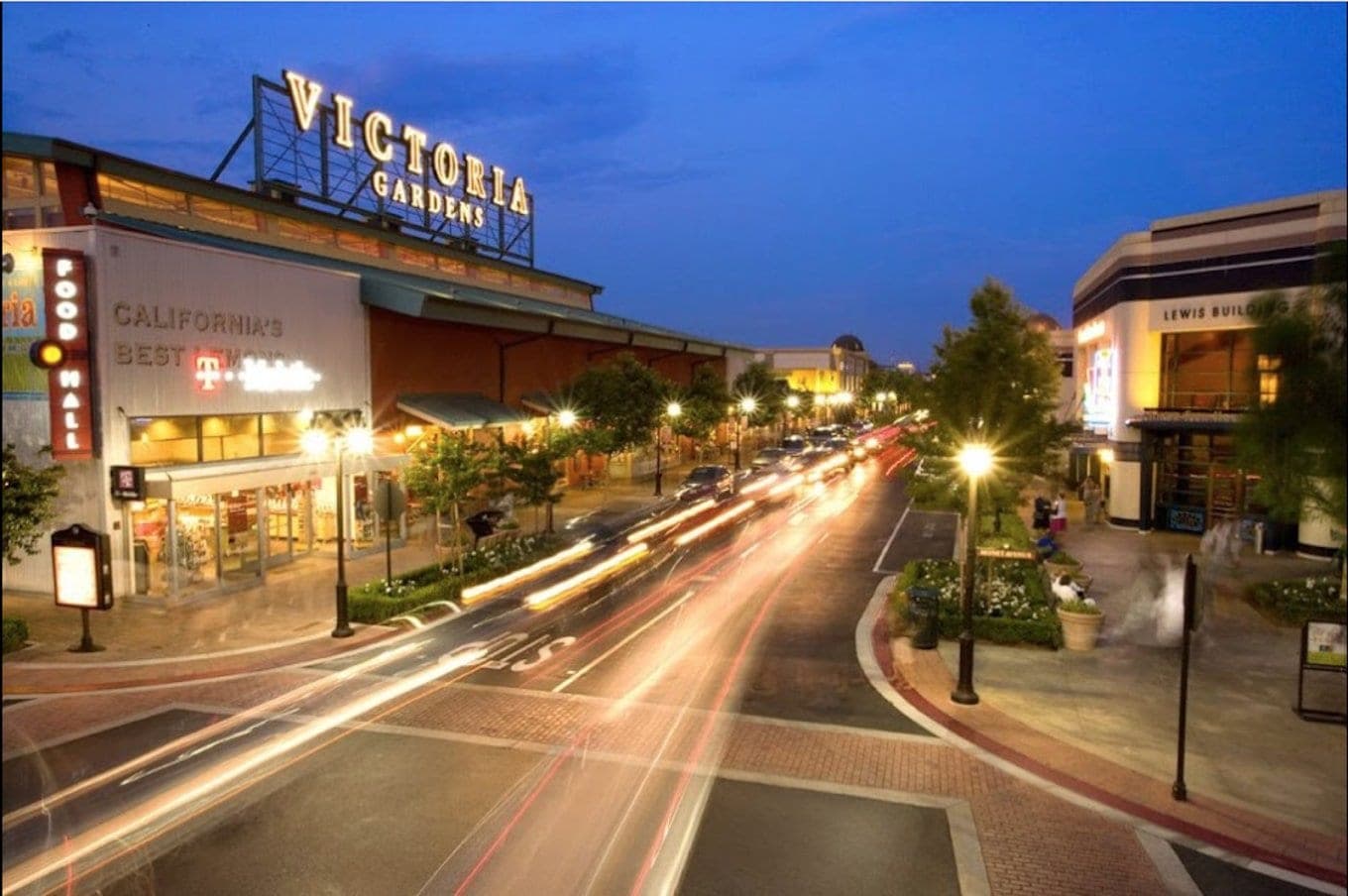 The Victoria Gardens sign above the main street at dusk, car lights streaking past lit storefronts